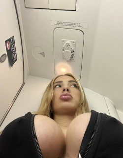 Blonde woman with a curvy build lies on a white surface in a tight black top, looking upward with lips puckered.