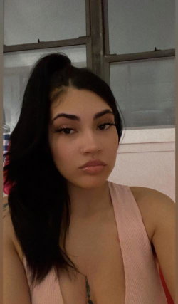 Brunette woman with long dark hair in a high ponytail, wearing a light pink halter top, taking a close-up selfie