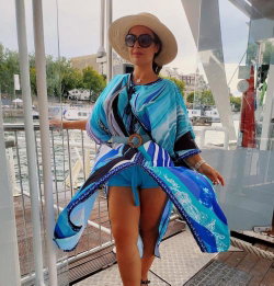 A woman in a blue patterned cover-up and straw hat poses on a boat deck, wearing sunglasses and shorts with a city