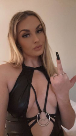 Blonde woman in a black strappy top with cutouts, making a hand gesture indoors.
