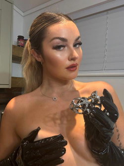 Blonde woman with ponytail, wearing black latex gloves, topless, holding a metallic object in a kitchen setting.