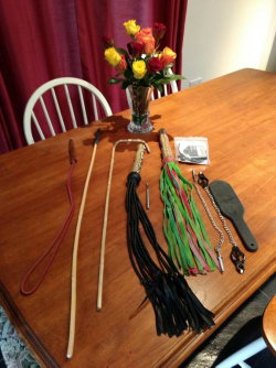 Various BDSM implements including whips, flogger, paddle, and chains arranged on a wooden table with a vase of roses in