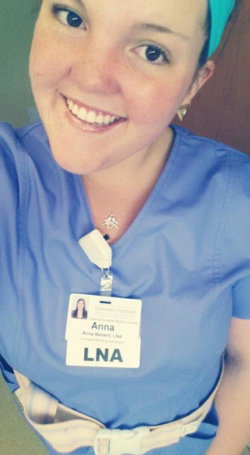 A smiling woman in blue medical scrubs with a name badge, wearing a teal headband and a stethoscope around her neck.