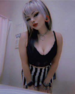 A woman with silver and black hair, bold makeup, and a black lace top poses in a bathroom mirror selfie, wearing