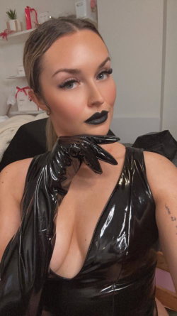 A woman with dark lipstick and dramatic makeup wears a shiny black latex top and long gloves, posing indoors with one