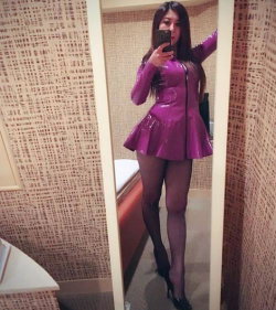 Brunette woman in a shiny purple mini dress, black stockings, and heels takes a mirror selfie in a patterned room.