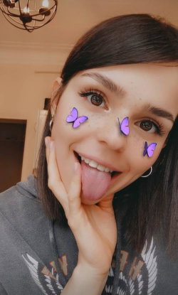 Brunette woman with freckles and butterfly face filters sticks out her tongue in a playful selfie indoors.