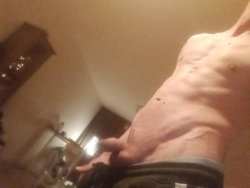 A shirtless man with a muscular torso is shown in a dimly lit room, holding the camera for a close-up selfie.