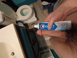 A person's hand holds a tube of Crest toothpaste in a bathroom with a toilet and sink visible.