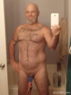Bald man with a gray beard and chest tattoos takes a selfie in a bathroom, holding a phone, wearing an American