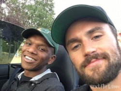 Two men smiling in a car, one wearing a green cap and the other with a beard and green cap, both looking at the camera.