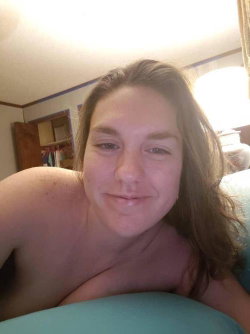 A woman with shoulder-length brown hair lies on a bed, bare-chested, smiling softly at the camera in a warmly lit