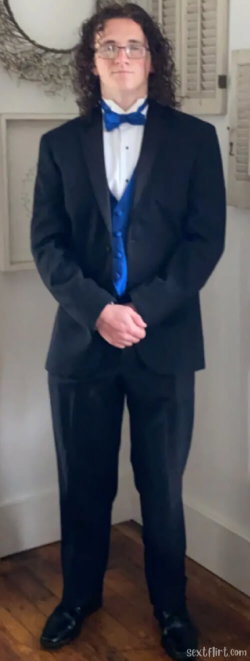 Person with curly hair and glasses wearing a black tuxedo with a blue bow tie and vest, standing indoors on wooden