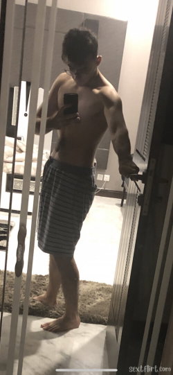 Shirtless muscular man with glasses takes mirror selfie in striped shorts, standing barefoot in a modern bedroom.