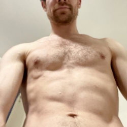 Shirtless man with short beard and chest hair, showing toned upper body in a close-up selfie indoors.
