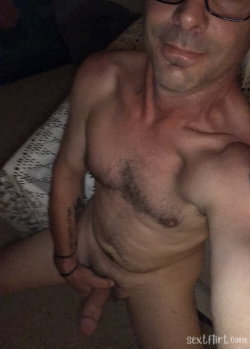 Shirtless man with glasses and chest hair, holding his penis while sitting on a patterned blanket in a dimly lit room.