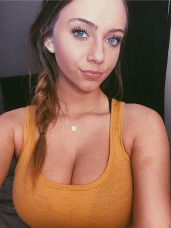 A woman with blue eyes and long brown hair in a braid wears a mustard yellow tank top, posing indoors with soft