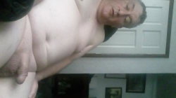 A shirtless man with a large belly lies on his back indoors, partially covered by dark fabric, in a dimly lit room with