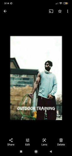 A muscular man with a beard wears a light blue long-sleeve shirt and dark pants, holding a green hose outdoors near a