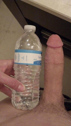 A hand holds a water bottle next to an erect penis, with the focus on the genitalia and the bottle for size comparison.