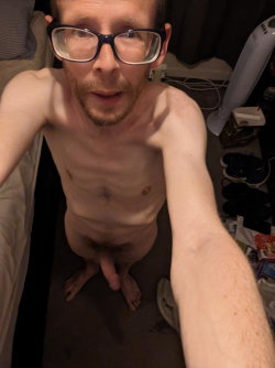 Shirtless man with glasses and short beard taking a selfie in a dimly lit room, standing barefoot with arm extended.