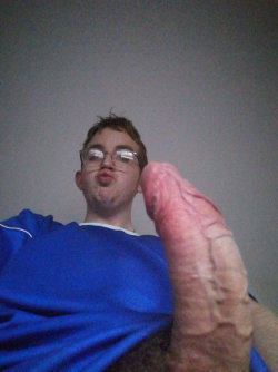 A young man with glasses makes a playful face while holding his erect penis close to the camera in a dimly lit room.