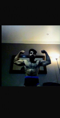 A muscular man with a beard flexes his arms in front of a mirror, wearing dark shorts with a blue waistband.
