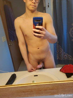 Shirtless man with glasses taking a mirror selfie in a bathroom, holding phone, partially covering genital area.
