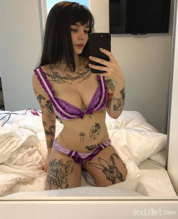 A woman with dark hair and multiple tattoos wears purple lace lingerie while taking a mirror selfie on a bed.