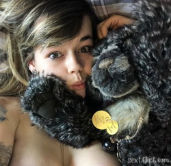 A young woman with light brown hair and visible tattoos lies topless, hugging a large black furry bear plushie close to