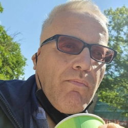 A man with short blond hair and sunglasses takes a selfie outdoors, holding a green cup, wearing a dark jacket over a
