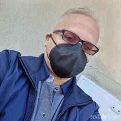 A man with short gray hair wearing sunglasses and a black face mask, dressed in a blue jacket over a collared shirt,