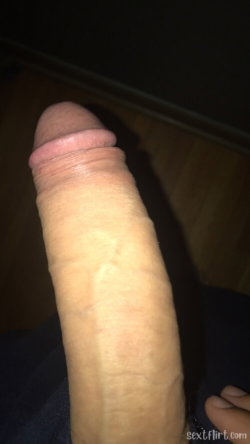 Close-up of an erect penis with visible skin texture, held against a dark background with wooden flooring.
