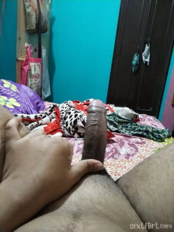 A person's hand holds a brown dildo on a patterned bedspread in a room with teal walls and scattered clothing.