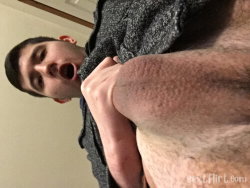 A man with short dark hair, wearing a gray sweater, holds his leg close to his face in a selfie-style shot indoors.