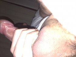 Close-up of a person's hand gripping a penis, with visible pubic hair and part of a white shirt in a dimly lit indoor
