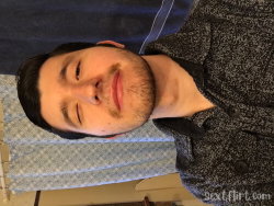 A man with short dark hair and a beard, wearing a patterned button-up shirt, smiles slightly in a close-up selfie