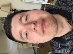 Close-up selfie of a man with dark hair and stubble, wearing a gray patterned shirt, indoors with furniture in the