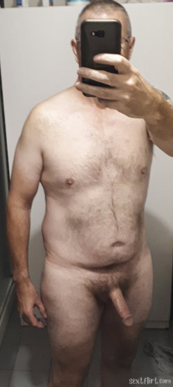 Shirtless man with visible chest and pubic hair taking a mirror selfie in a tiled room, holding phone to cover face.