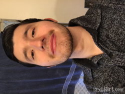 A man with short dark hair and a light beard lies on a bed, wearing a patterned button-up shirt, looking at the camera