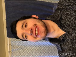 A smiling man with short dark hair and a beard, wearing a patterned button-up shirt, lying on a bed with a blue towel