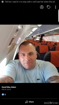 A man with short hair and visible arm tattoos takes a selfie in an airplane cabin, wearing a light blue t-shirt.