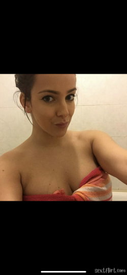 A woman with dark hair tied back, wearing a striped towel around her torso, looks at the camera in a tiled bathroom