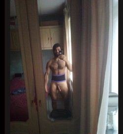 A shirtless man with a beard takes a mirror selfie in a bedroom, wearing purple-trimmed underwear, with his genitals