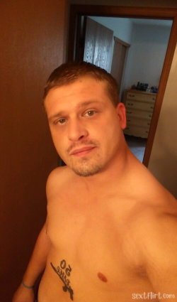 Shirtless man with short hair and stubble, showing chest tattoo, standing indoors near a doorway.