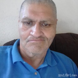 An older man with short gray hair and a blue polo shirt, sitting indoors against a plain wall.