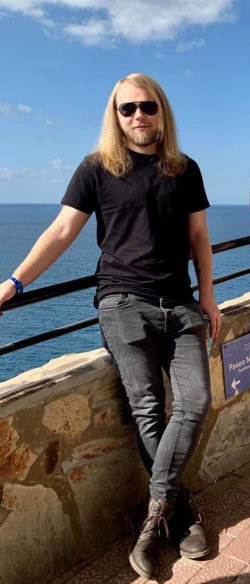 A man with long blonde hair and a beard wears a black t-shirt and dark jeans, leaning against a stone railing by the