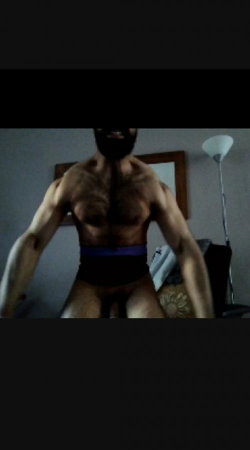 Muscular man with beard, wearing dark underwear with purple waistband, standing indoors in dim lighting.