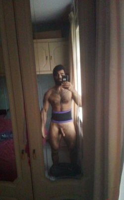 A muscular man with a beard takes a mirror selfie in a room, wearing only a purple and black waistband, showing his