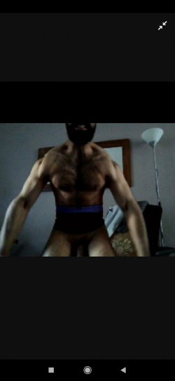 Muscular man with beard, shirtless, wearing dark underwear with purple waistband, standing indoors near lamp and framed